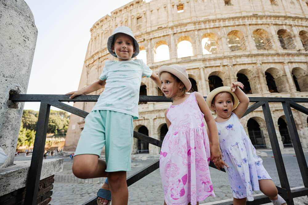 Three children in sunhats pose for a photo in front of the Colosseum in Rome