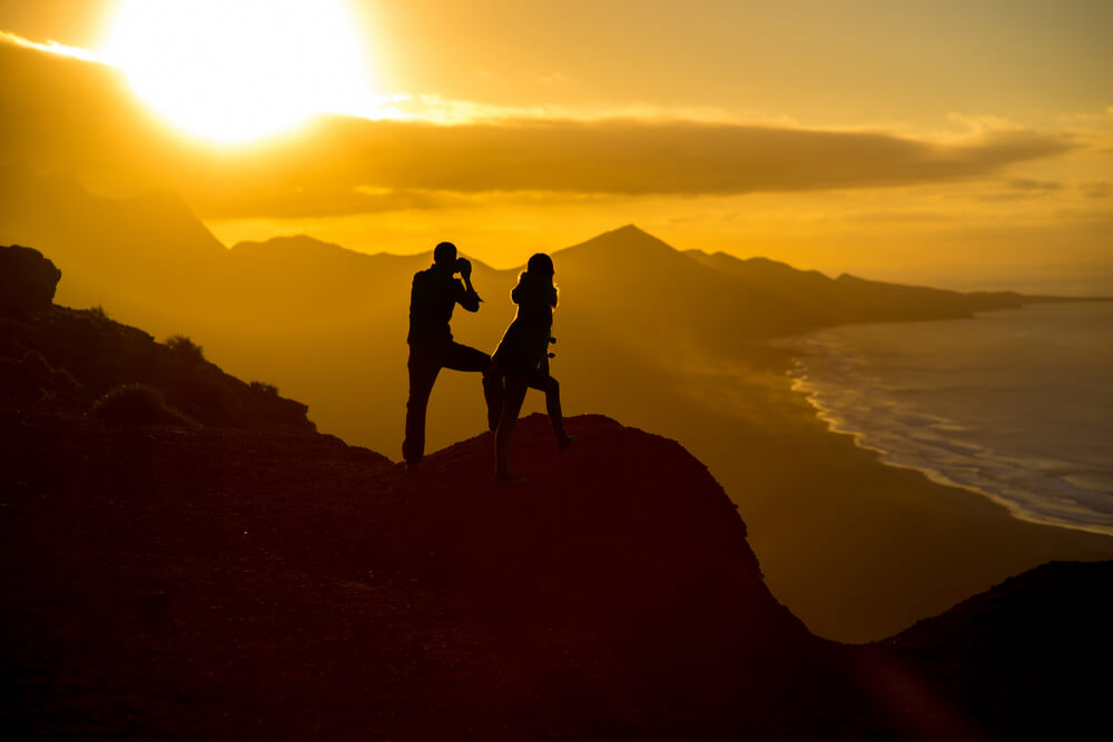 A couple silhouetted against the setting sun take photos in mountain scenery overlooking a beach