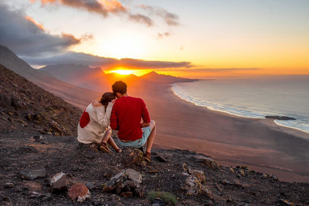 A couple watch the sun set behind volcanic mountains above a huge deserted beach