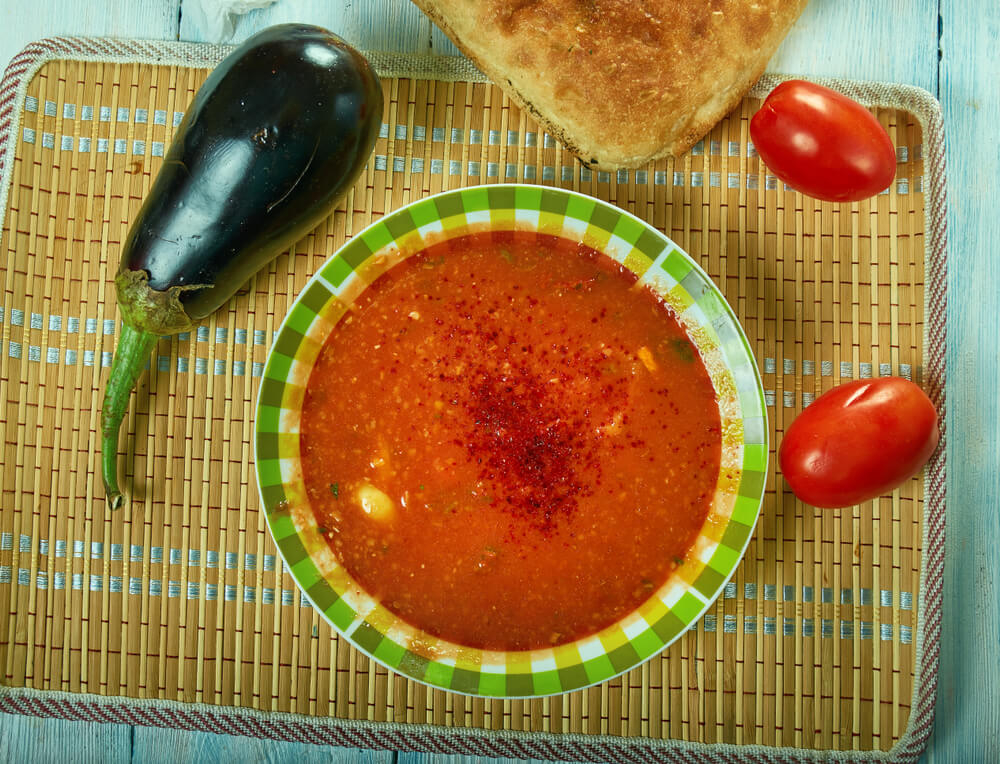 A bowl of spicy Bulgarian tripe soup flanked by an aubergine, tomatoes and bread