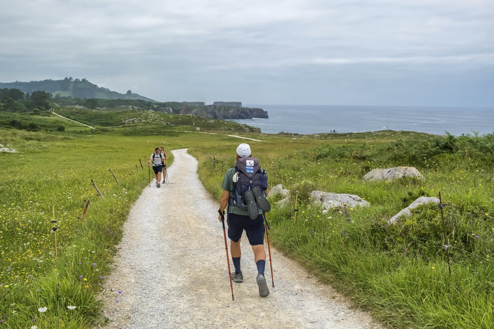 Hikers walk a dirt path amid green fields on the north coast of Spain