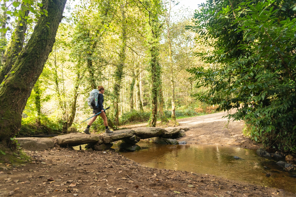Camino de Santiago routes: a pilgrim crosses a stone bridge over a stream amid woodland in Galicia