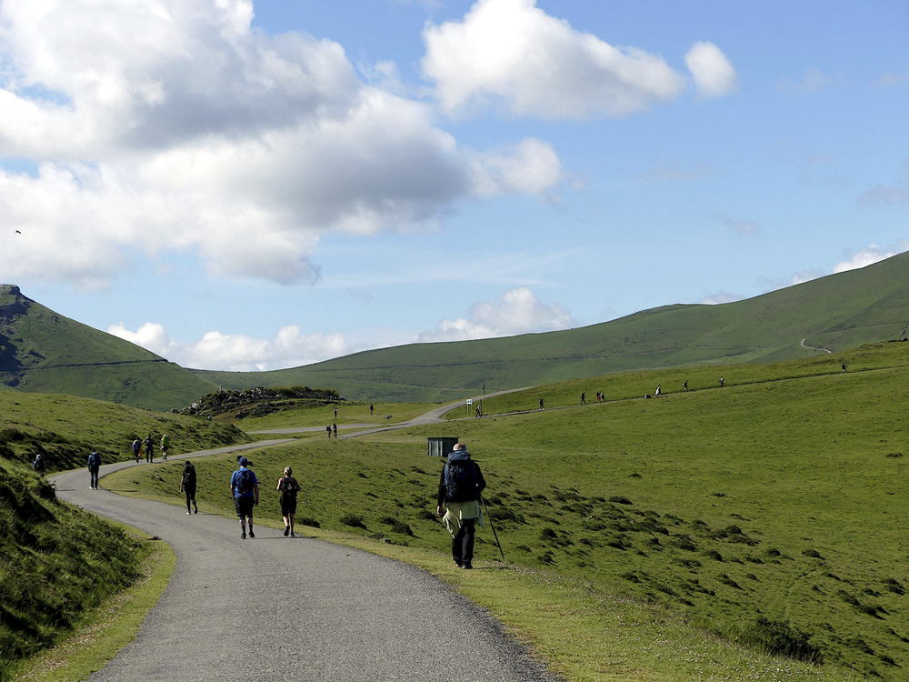 Hikers head up a rolling green valley into the Pyrenees under a blue sky and white clouds