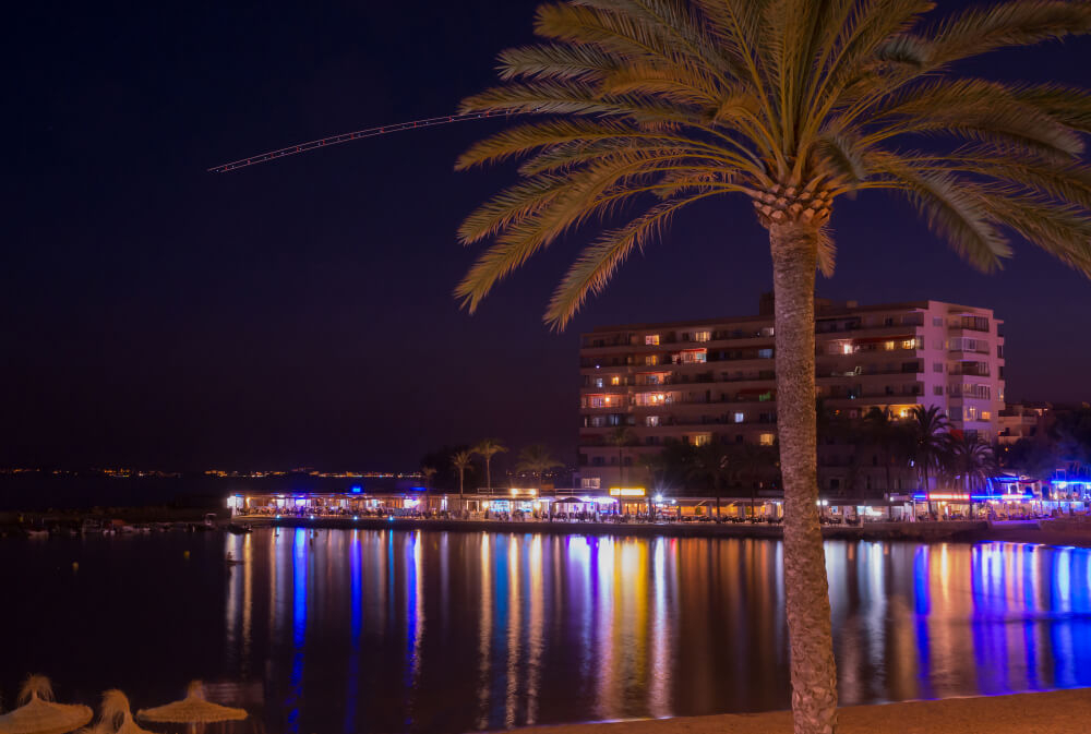 Distant brightly lit bars reflect in the water in night-time Magaluf, a palm is in the foreground