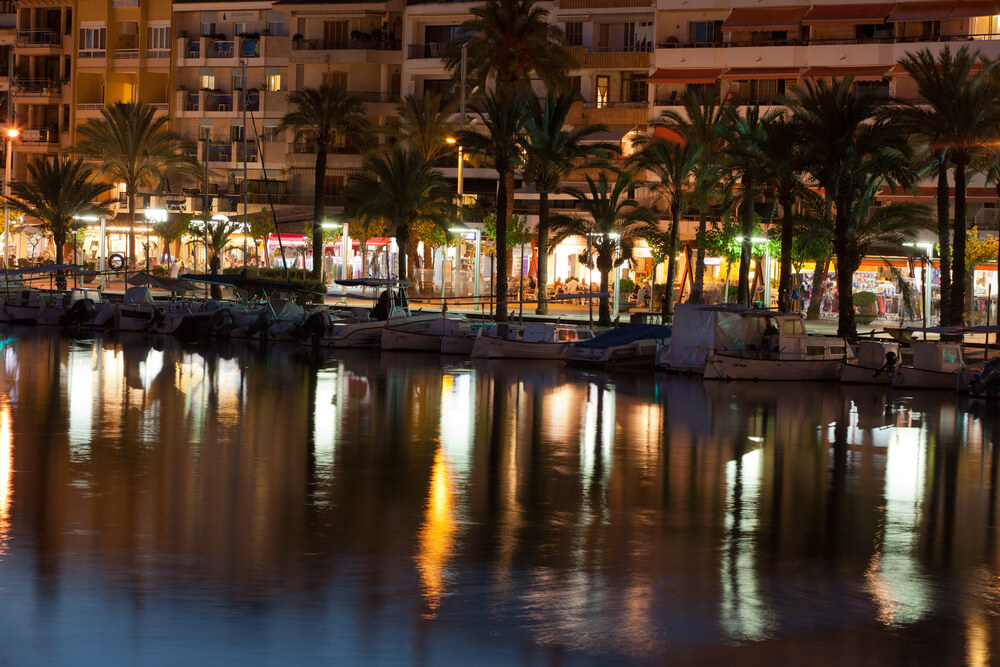 Bars, boats and palm trees shimmer in the water on Alcudia waterfront by night