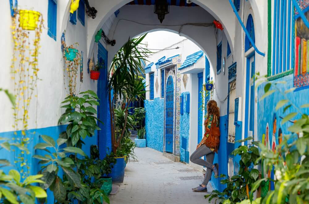 A woman leans against an arch in a Tangier lane painted blue and white and lined with plants
