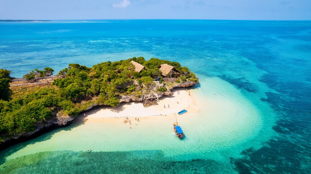 A bird’s eye view of a boat resting near the shore of a tropical beach backed by low cliffs and huts